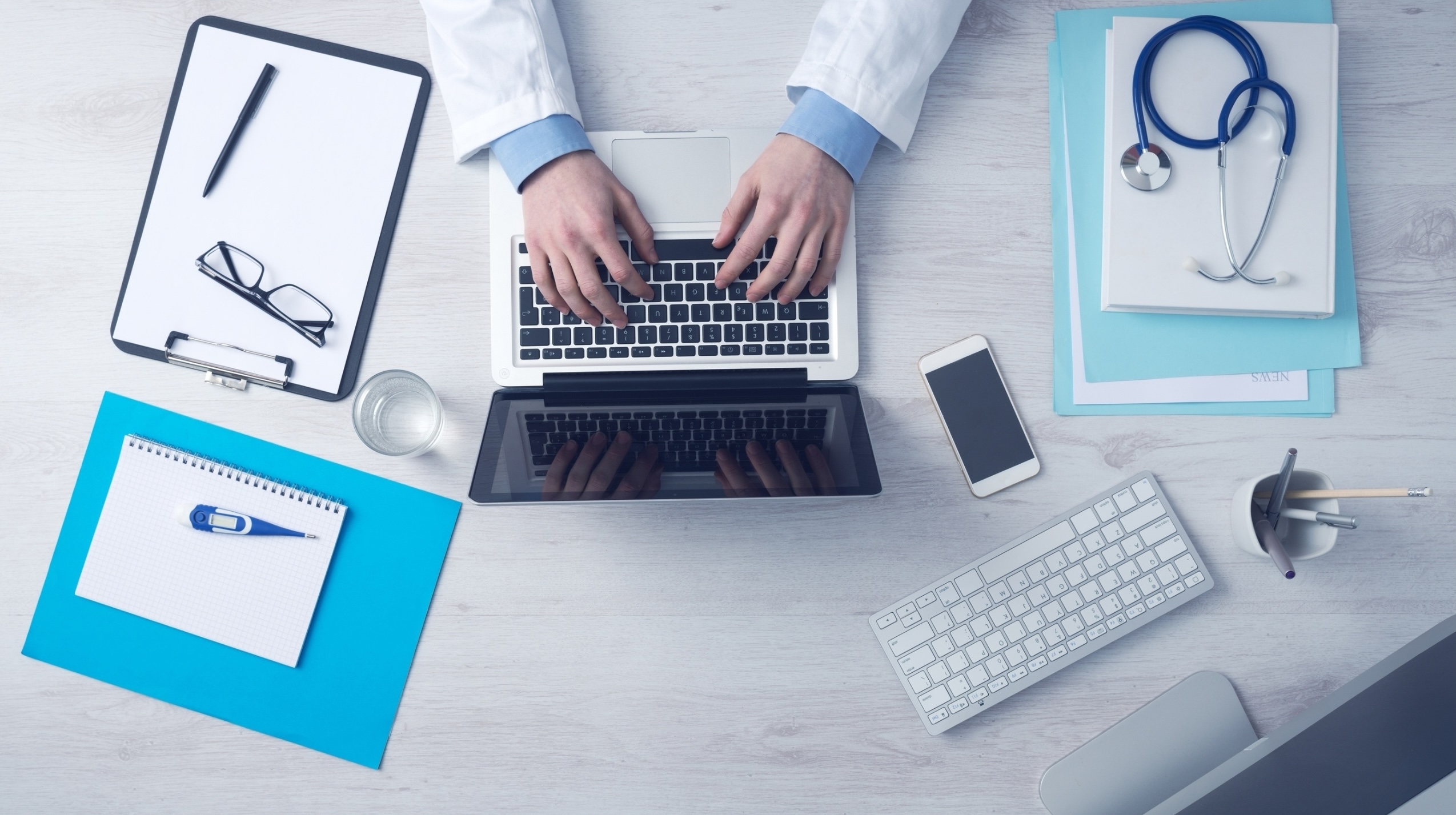 Top down view of doctor using his laptop with a mobile phone and desktop computer nearby.